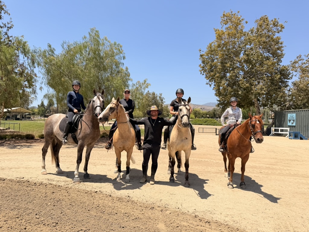Four riders on horseback lined up in an arena with their trainer standing between them
